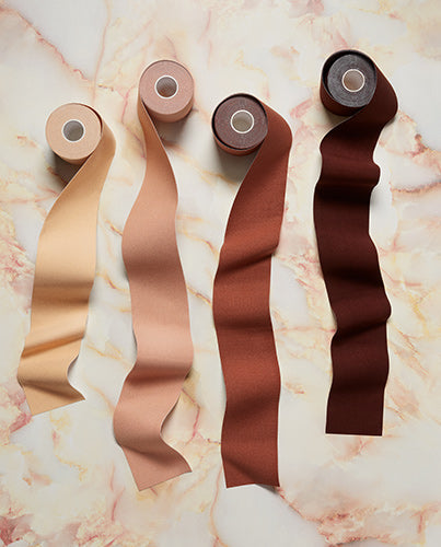 Four rolls of adhesive boob tape in diverse skin tones cascade down a pink marble background.