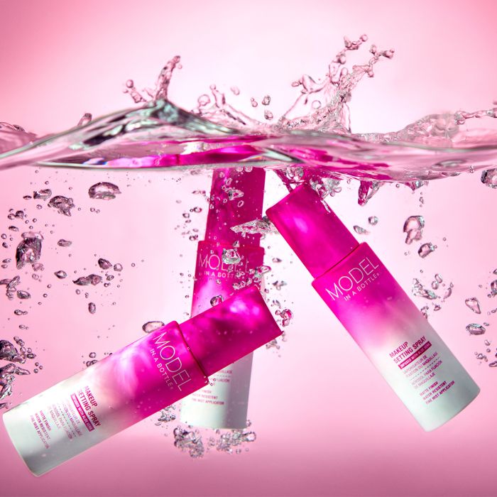 Three bottles of Model in a Bottle Makeup Setting Spray submerge in splashing pink water to showcase their water-resistant and matte finish formula.