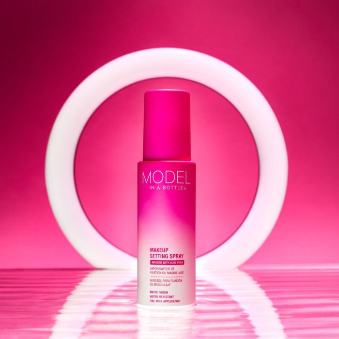 A bottle of Model In A Bottle matte finish makeup setting spray stands centered against a glowing white ring light on a vibrant pink background.