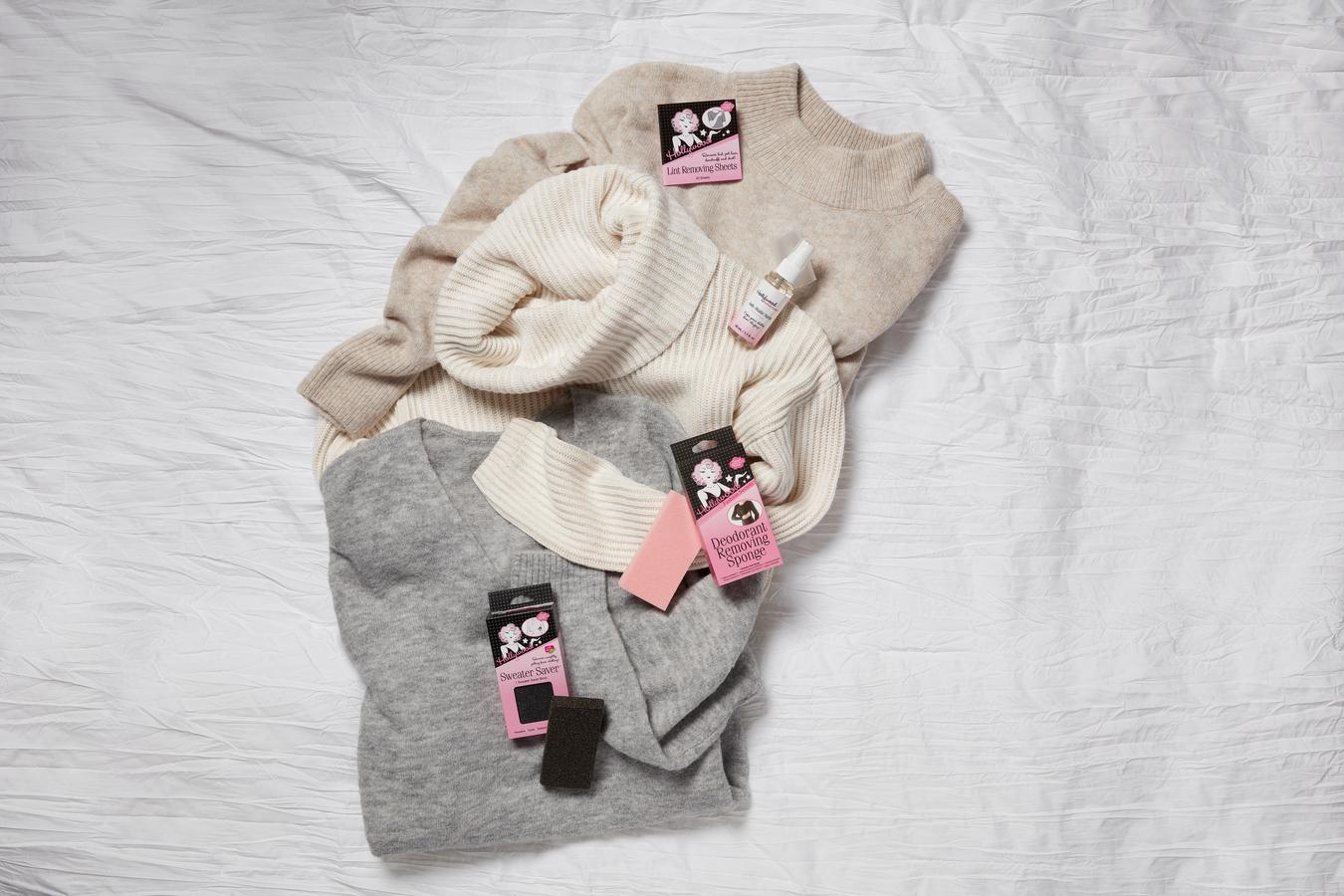 Three neutral-toned knit sweaters rest on a white textured surface alongside essential garment care accessories like a pilling stone and lint removal sheets.