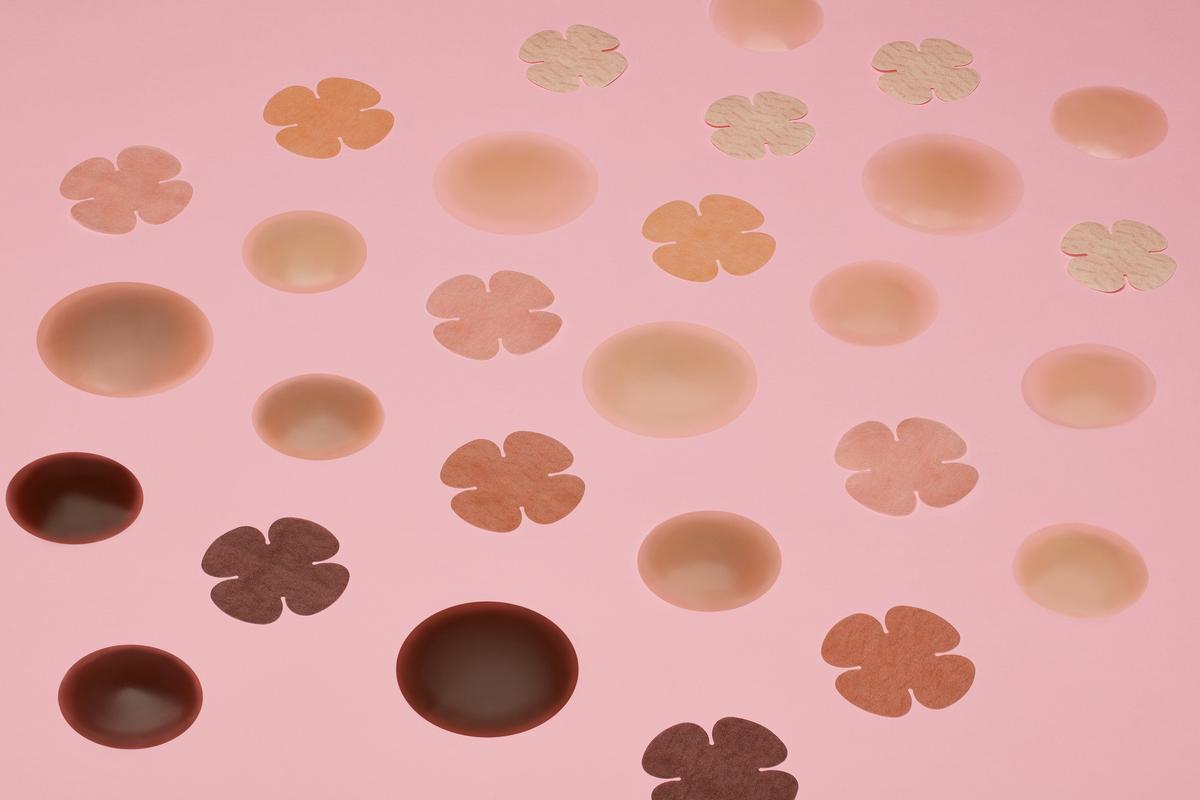 A collection of silicone nipple covers and flower-shaped adhesive pasties in diverse skin tones rests on a vibrant pink background.