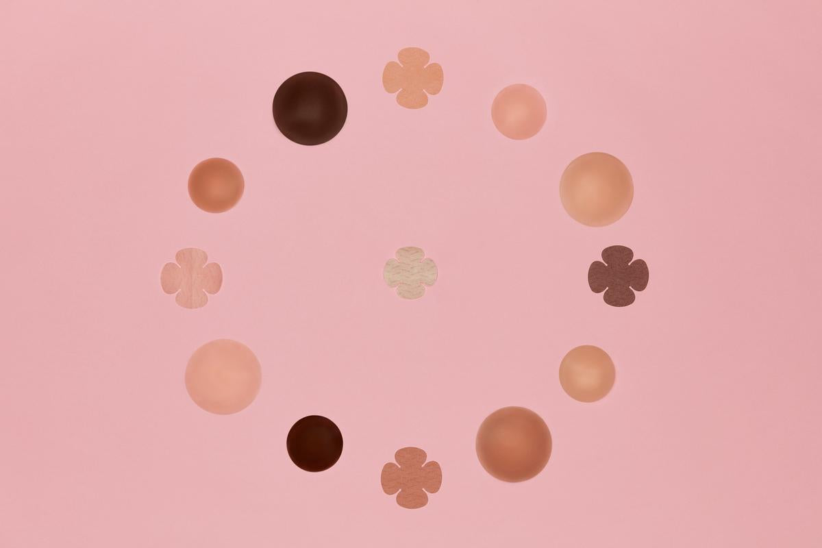 Diverse skin-toned nipple covers and adhesive pasties in various shapes form a circular arrangement on a soft pink background.