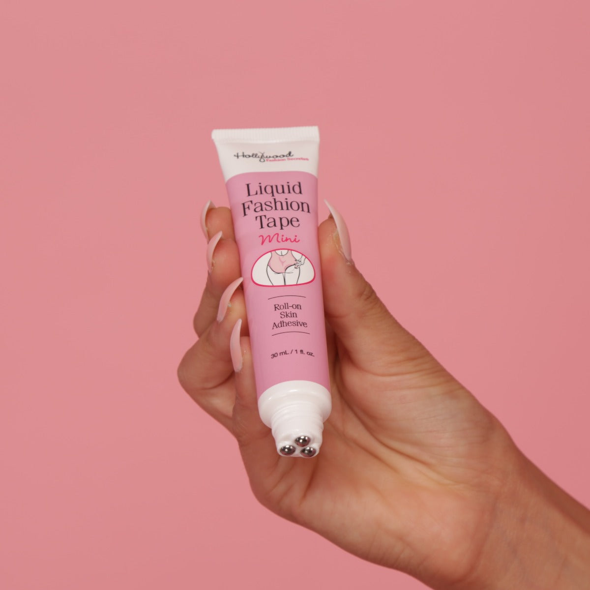 A hand holds a tube of Hollywood Fashion Secrets Liquid Fashion Tape Mini, showcasing the roll-on skin adhesive applicator against a pink background.