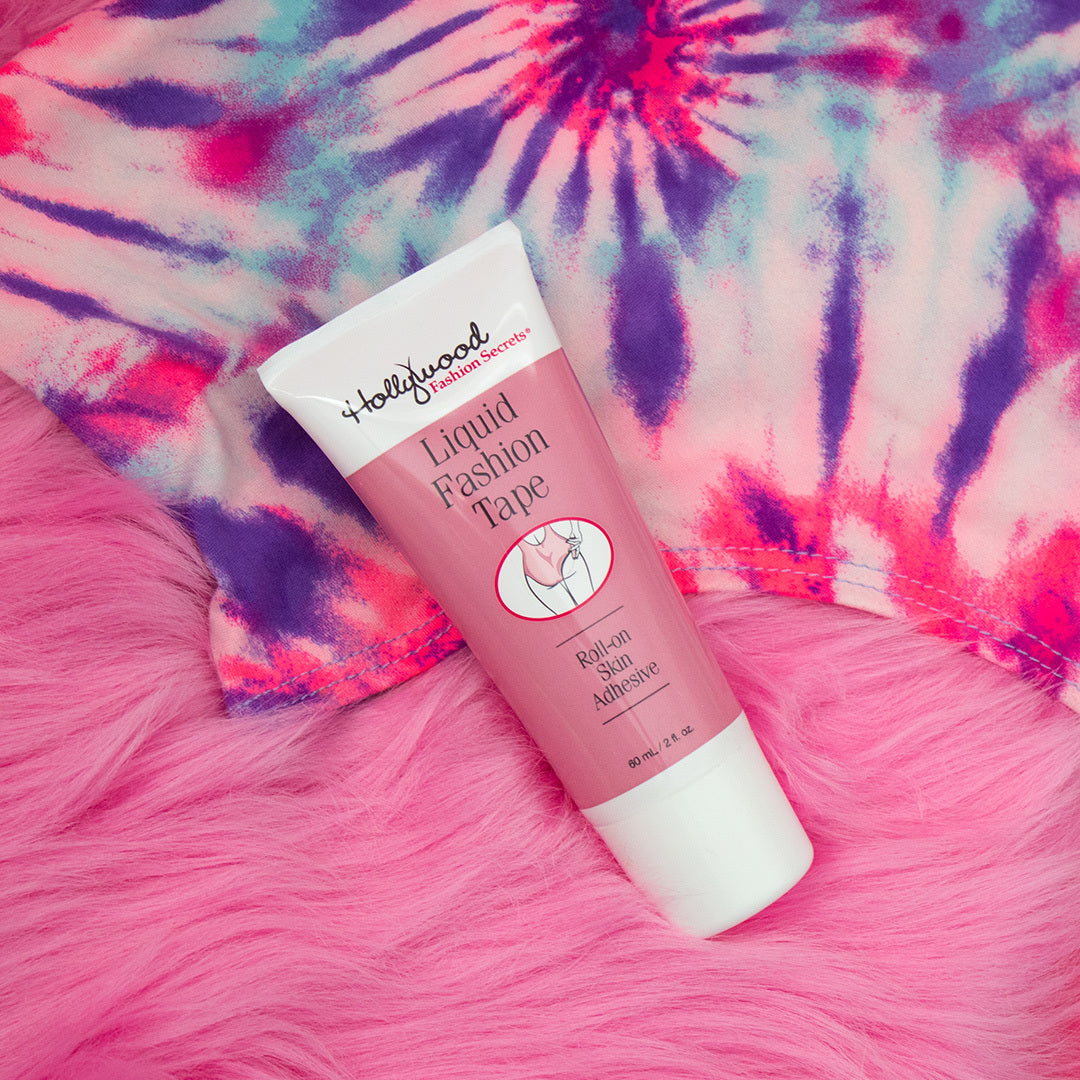 A tube of Hollywood Fashion Secrets Liquid Fashion Tape roll-on skin adhesive rests on a plush pink faux fur surface beside a vibrant tie-dye top.
