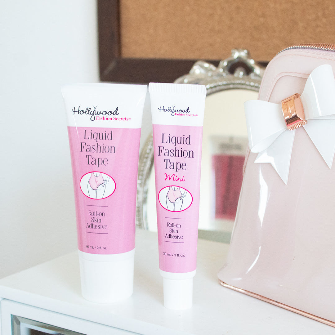 Two tubes of Hollywood Fashion Secrets Liquid Fashion Tape roll-on skin adhesive stand on a vanity beside a pink makeup bag with a white bow.