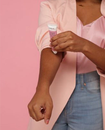 A woman in a pink blazer applies liquid fashion tape from a small tube to her inner arm against a vibrant pink background.