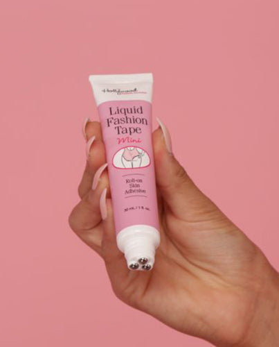 A hand with manicured nails displays a tube of Hollywood Fashion Secrets Liquid Fashion Tape Mini, highlighting its triple-rollerball skin adhesive applicator against a pink background.