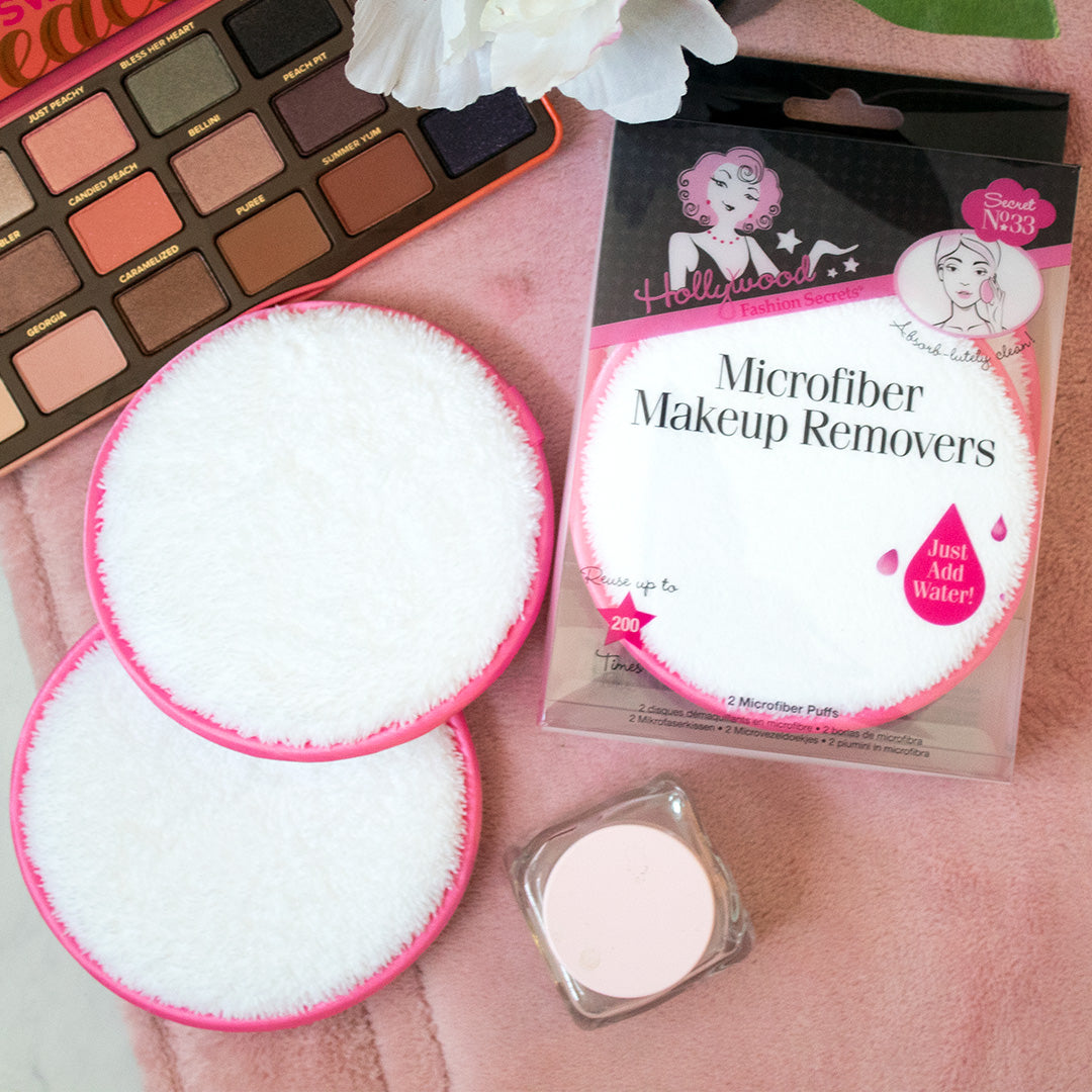 Two reusable white microfiber makeup remover pads with pink trim sit on a soft pink surface next to their packaging and a peach eyeshadow palette.