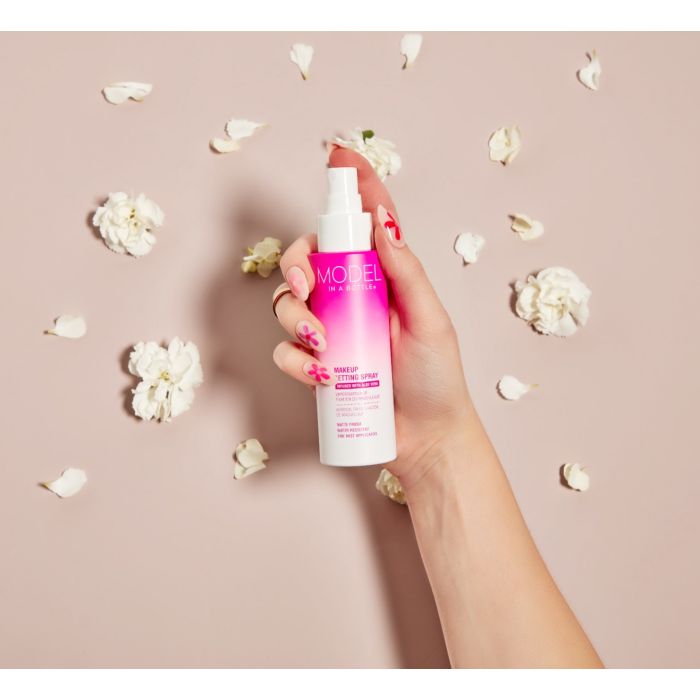 A manicured hand with pink floral nail art holds a bottle of Model in a Bottle makeup setting spray surrounded by scattered white flower petals.