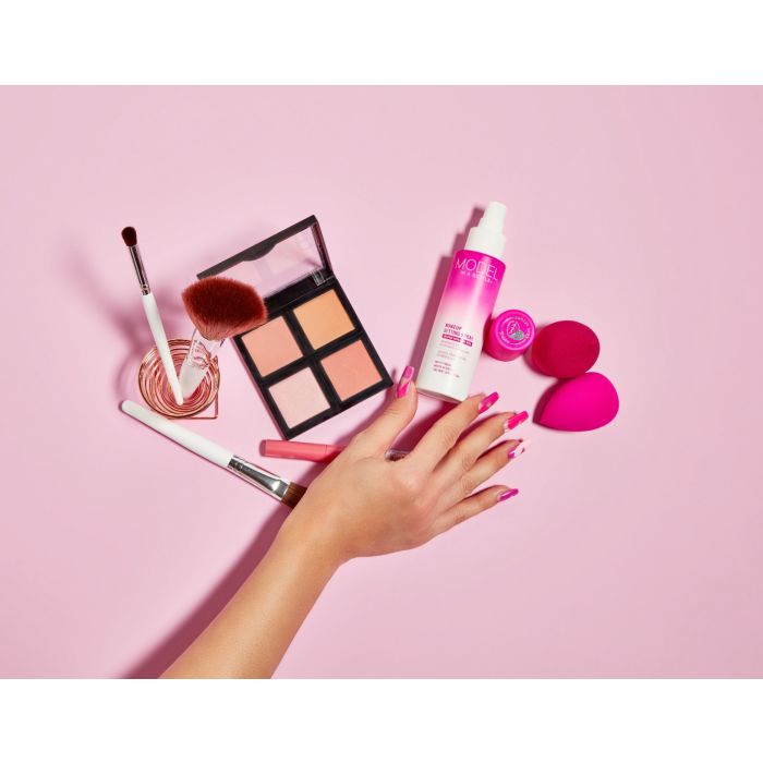 A hand with a pink patterned manicure reaches toward a collection of pink makeup products, including a face palette, setting spray, and makeup sponges, on a light pink background.