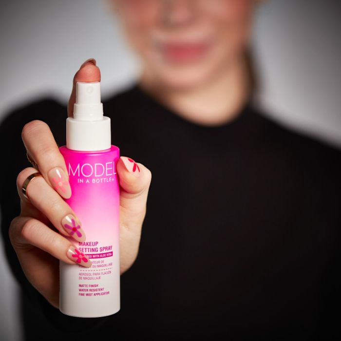 A hand with pink floral nail art holds a bottle of Model in a Bottle matte finish makeup setting spray.