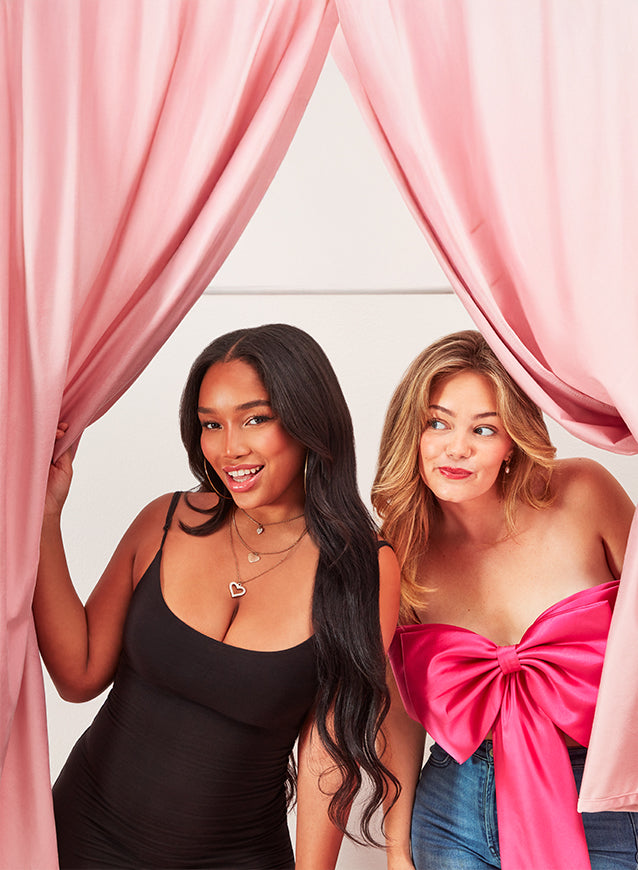 Two women standing behind pink curtains, one wearing a black top and the other a pink top with a bow.