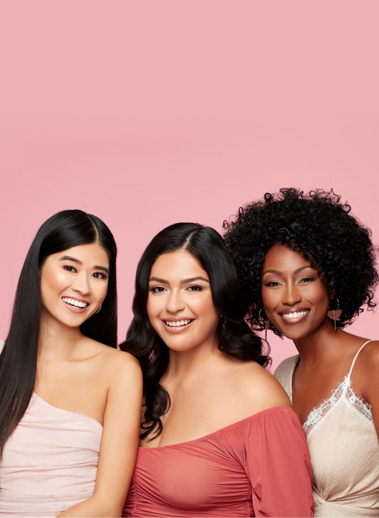 Diverse Women Smiling – Three diverse women in fashionable outfits posing against a pink background.