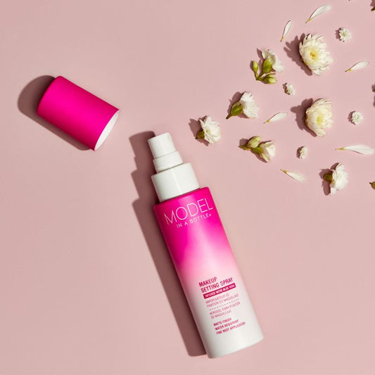 A bottle of Model in a Bottle matte makeup setting spray sits on a soft pink background surrounded by delicate white flowers.