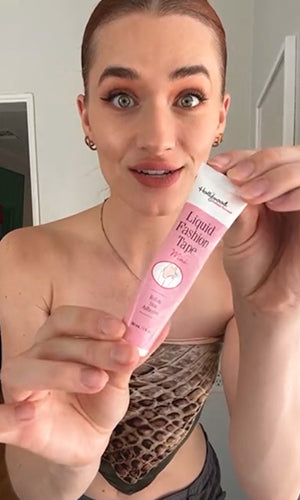 A woman holding up a pink tube of 'Liquid Fashion Tige' cosmetic product in front of her face, showing the packaging in a close-up shot.