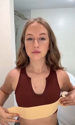 A woman with light brown hair is wearing a maroon sports bra and holding a beige underwire bra against her torso to show a comparison or alternative style. She is standing indoors against a white wall.