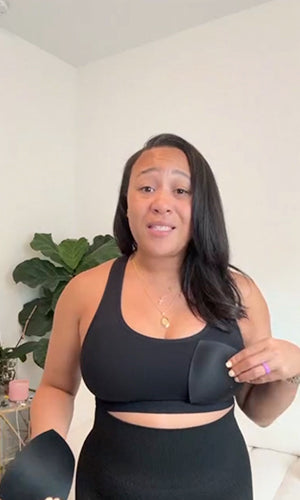 A woman wearing a black sports bra and matching high-waisted leggings, holding a black underwire bra in one hand and wearing a matching black bra cup on the other shoulder. She is standing in a brightly lit room with a white wall and a green potted plant in the background.