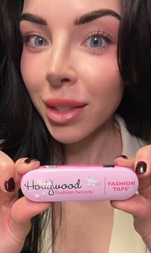 A close-up of a woman holding a pink compact case labeled 'Hollywood Fashion Secrets' and 'FASHION TAPE' in front of her face.