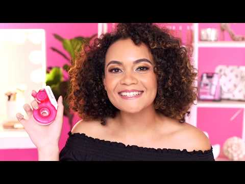 A smiling woman with curly brown hair showcases a pink donut-shaped eyelash tape dispenser against a vibrant pink background.