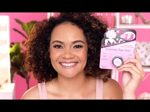 A smiling woman with curly brown hair holds a package of Hollywood Accessory Tape Dots in a brightly lit pink room.
