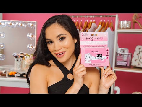 A smiling woman showcases a pink Hollywood Fashion Secrets Style Emergency Kit in front of her brightly lit fashion vanity.
