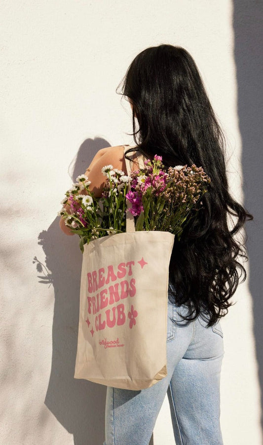 A woman with long, wavy dark hair carries a "Breast Friends Club" canvas tote bag overflowing with colorful flowers against a sunlit white wall.