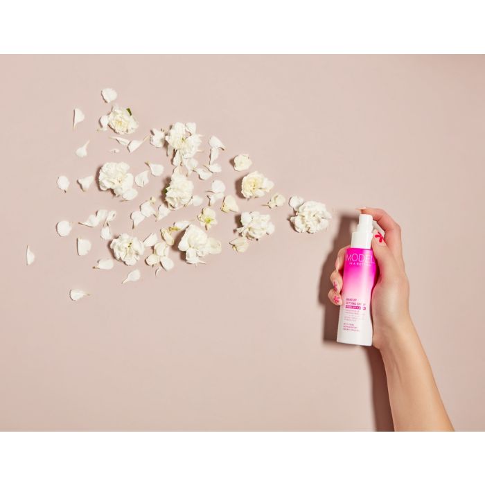 Hand spraying Model in a Bottle Makeup Setting Spray, with white flowers artistically arranged on a neutral background.