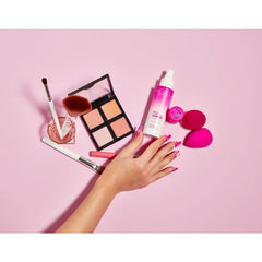 Flat lay of makeup essentials, including Model in a Bottle Setting Spray, brushes, beauty blenders, and a highlighter palette.